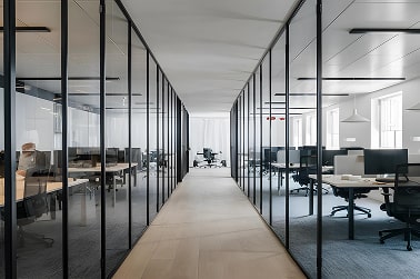 A modern office corridor with glass-walled rooms on either side, featuring desks and chairs.